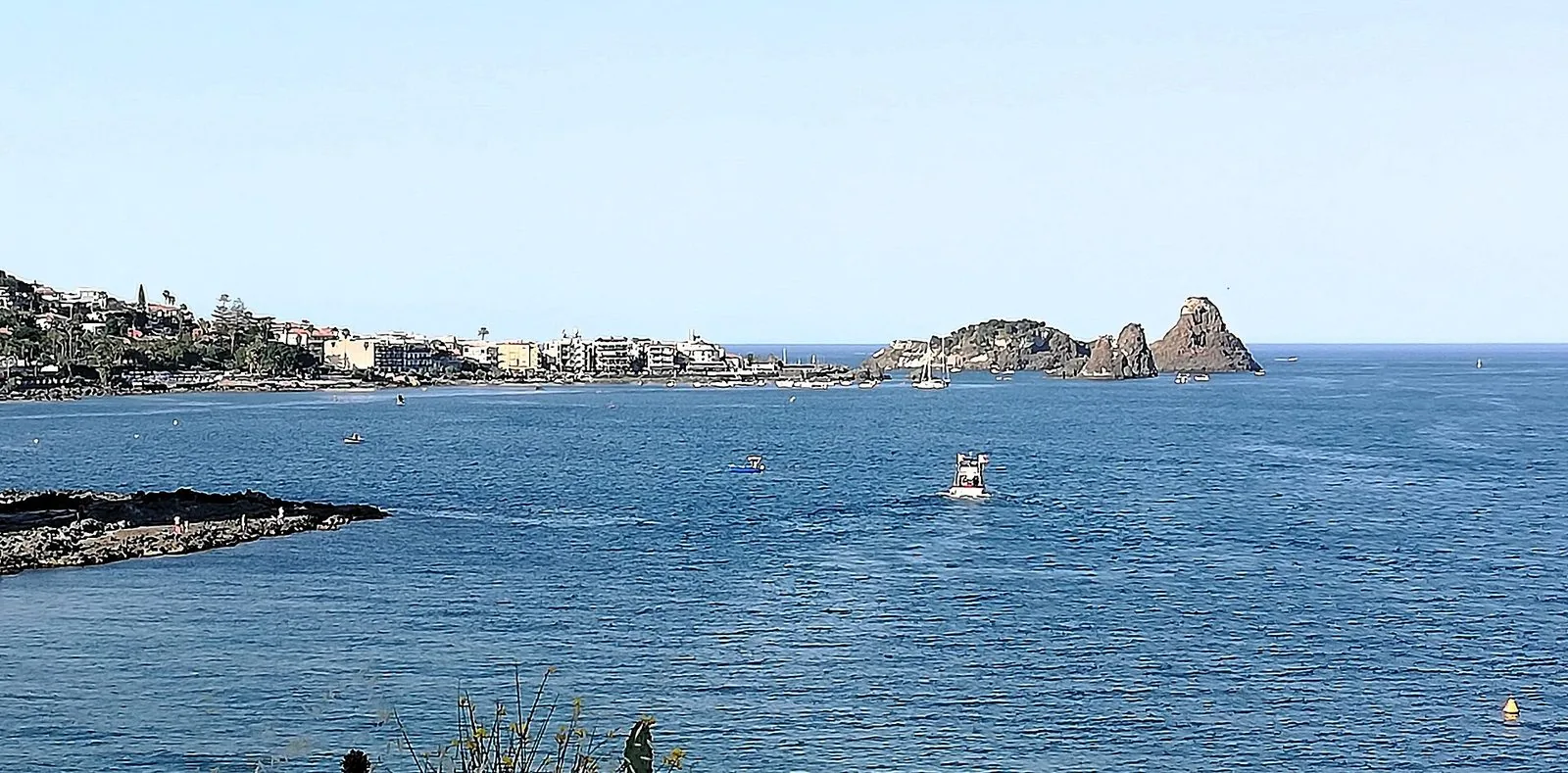 Il mare blu davanti ad Aci Trezza con il lungomare e le case del borgo a sinistra, l'Isola Lachea e i Faraglioni dei Ciclopi a destra e alcune barche in acqua