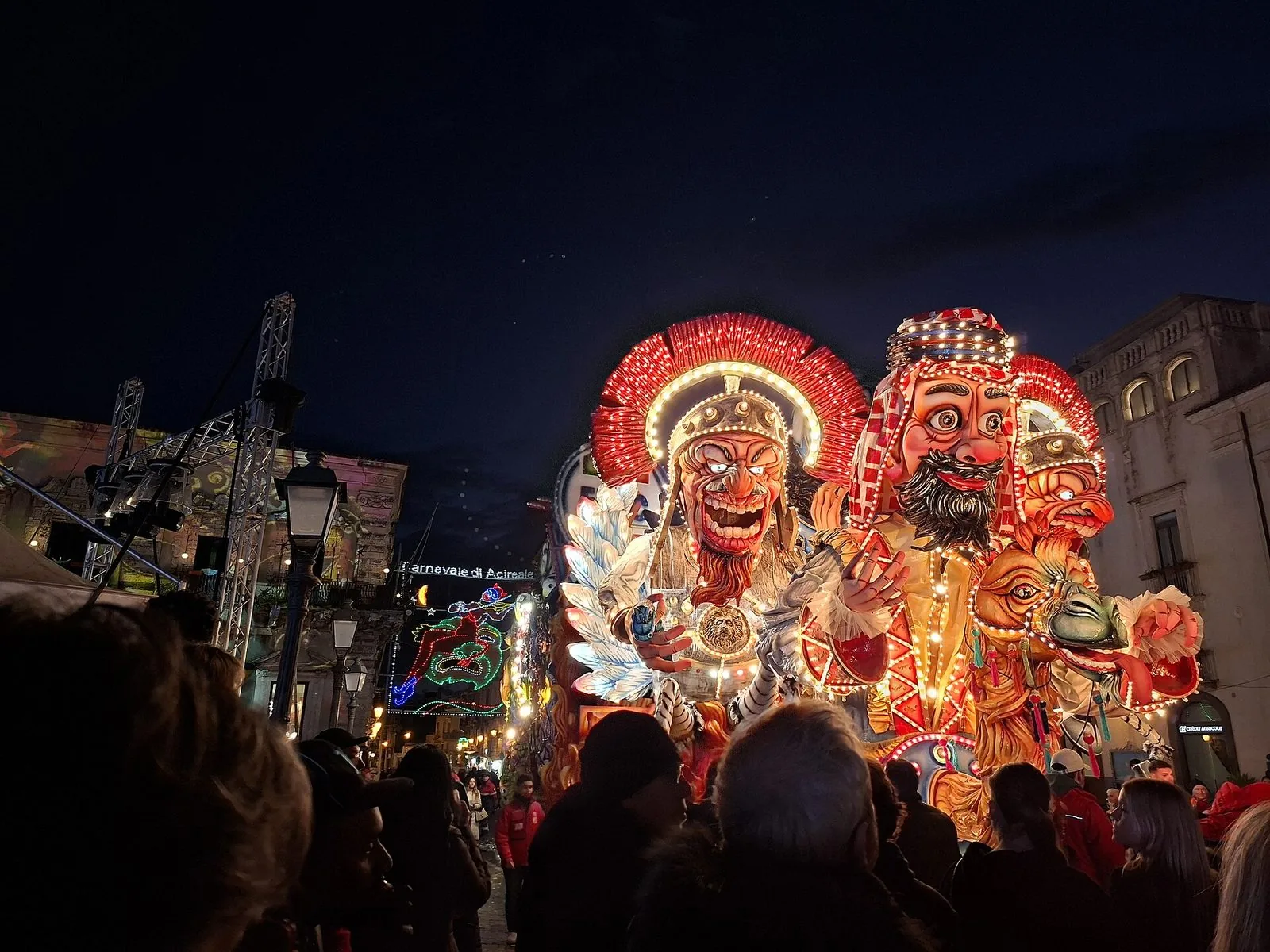 Un carro allegorico del Carnevale di Acireale di notte, con grandi teste caricaturali in cartapesta coperte di luci colorate, la folla di spalle in primo piano e una scritta luminosa del carnevale sulla strada