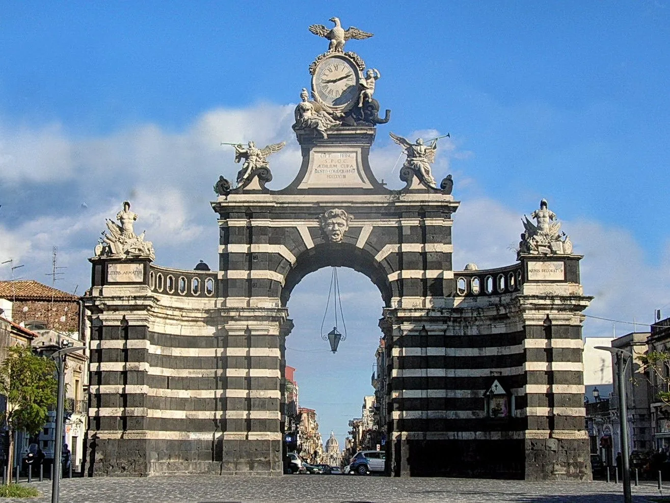 Porta Garibaldi a Catania, arco trionfale settecentesco a fasce alternate di pietra lavica nera e pietra bianca, sormontato da un orologio, un'aquila e statue di angeli, con il cielo azzurro sullo sfondo