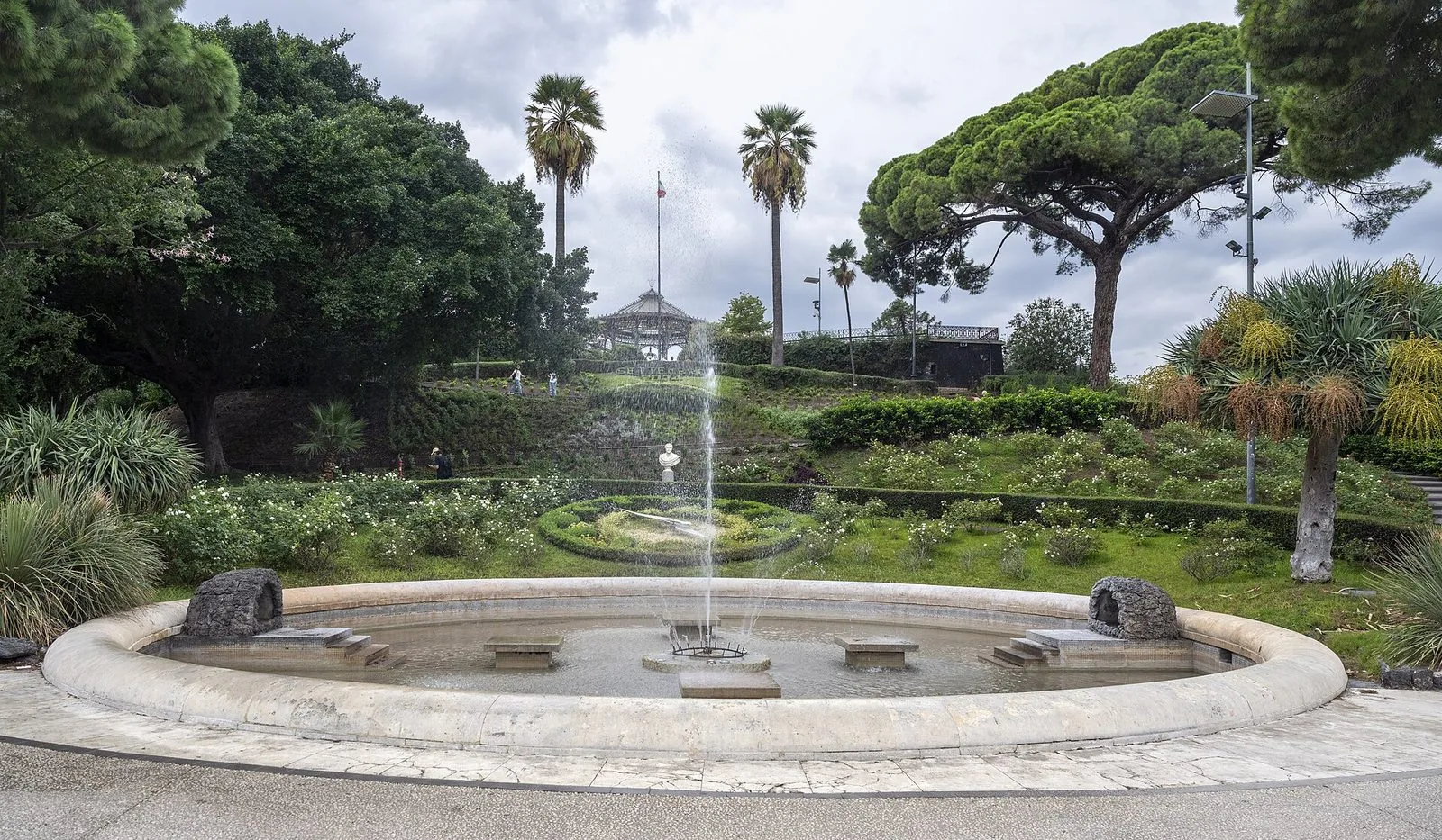 La fontana circolare con zampillo della Villa Bellini a Catania, circondata da aiuole di rose, palme e pini, con il chiosco della musica sulla collinetta sullo sfondo