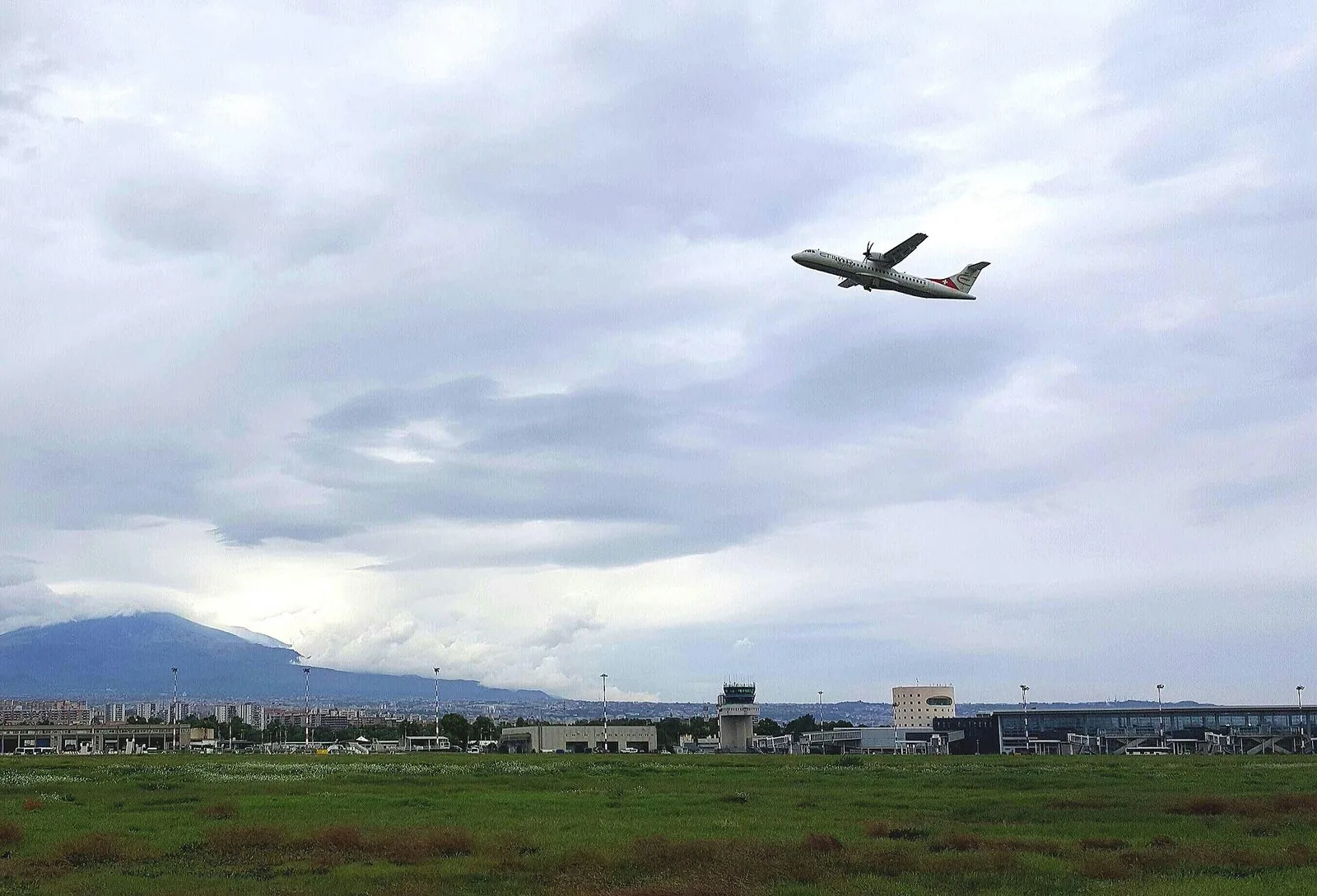 Un aereo a elica in decollo sopra il prato dell'aeroporto di Catania Fontanarossa, con la torre di controllo, i terminal e sullo sfondo l'Etna avvolto dalle nuvole