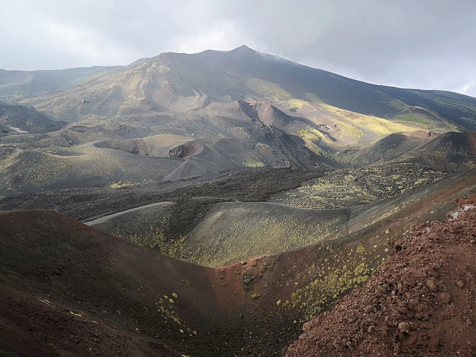 Il versante sud dell'Etna con i coni dei Crateri Silvestri in primo piano, sabbie vulcaniche rosse e nere, vecchie colate e la cima del vulcano sotto un cielo nuvoloso